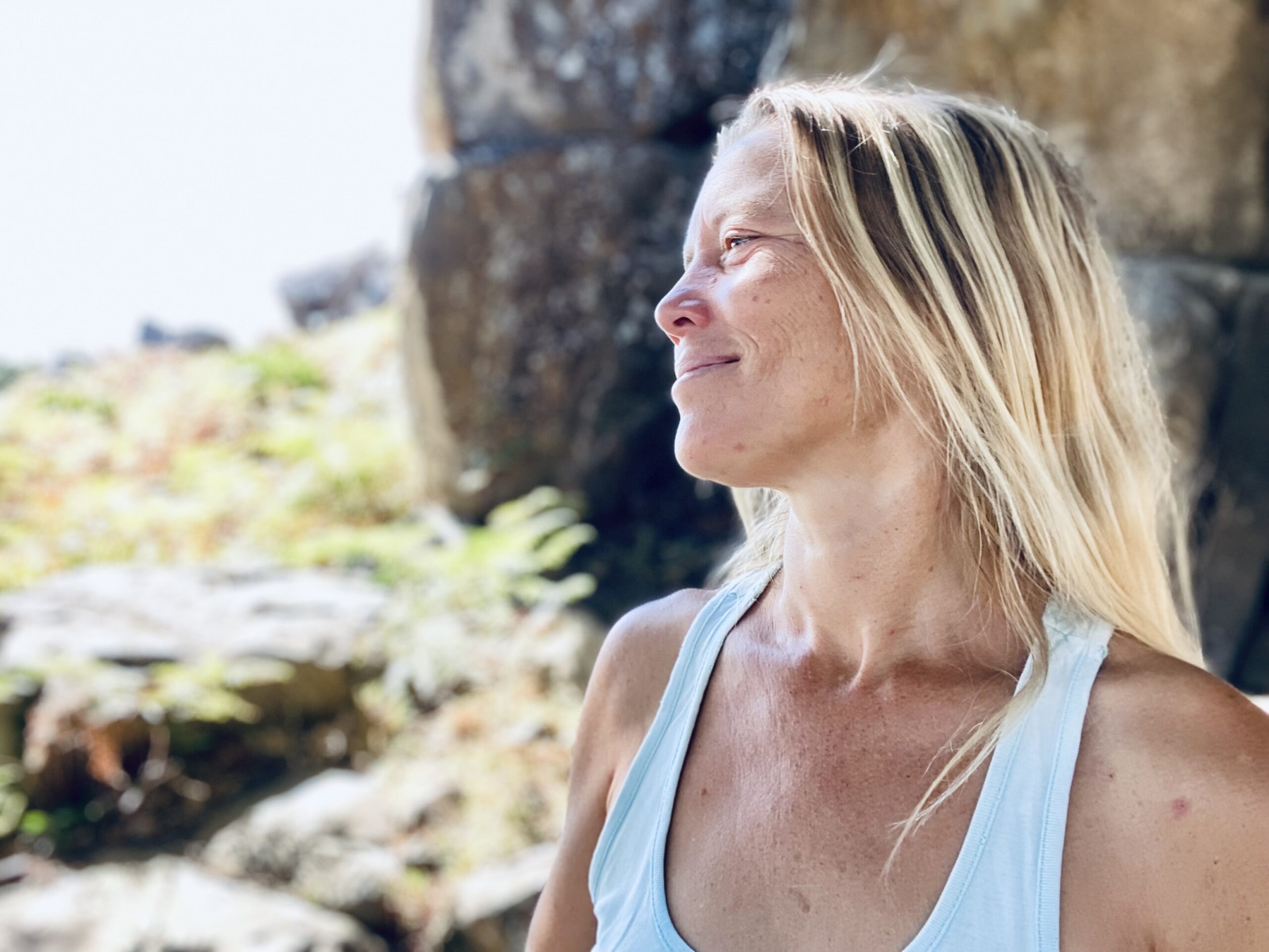 Auf dem Bild sieht man Astrid Schröder links von der Seite. Sie trägt ihre blonden langen Haare offen und locker nach hinten gelegt. Sie schaut verträumt, leicht nach rechts in die Landschaft . Im Hintergrund sieht man einen großen Felsen und Wiese. Die Sonne scheint. 
