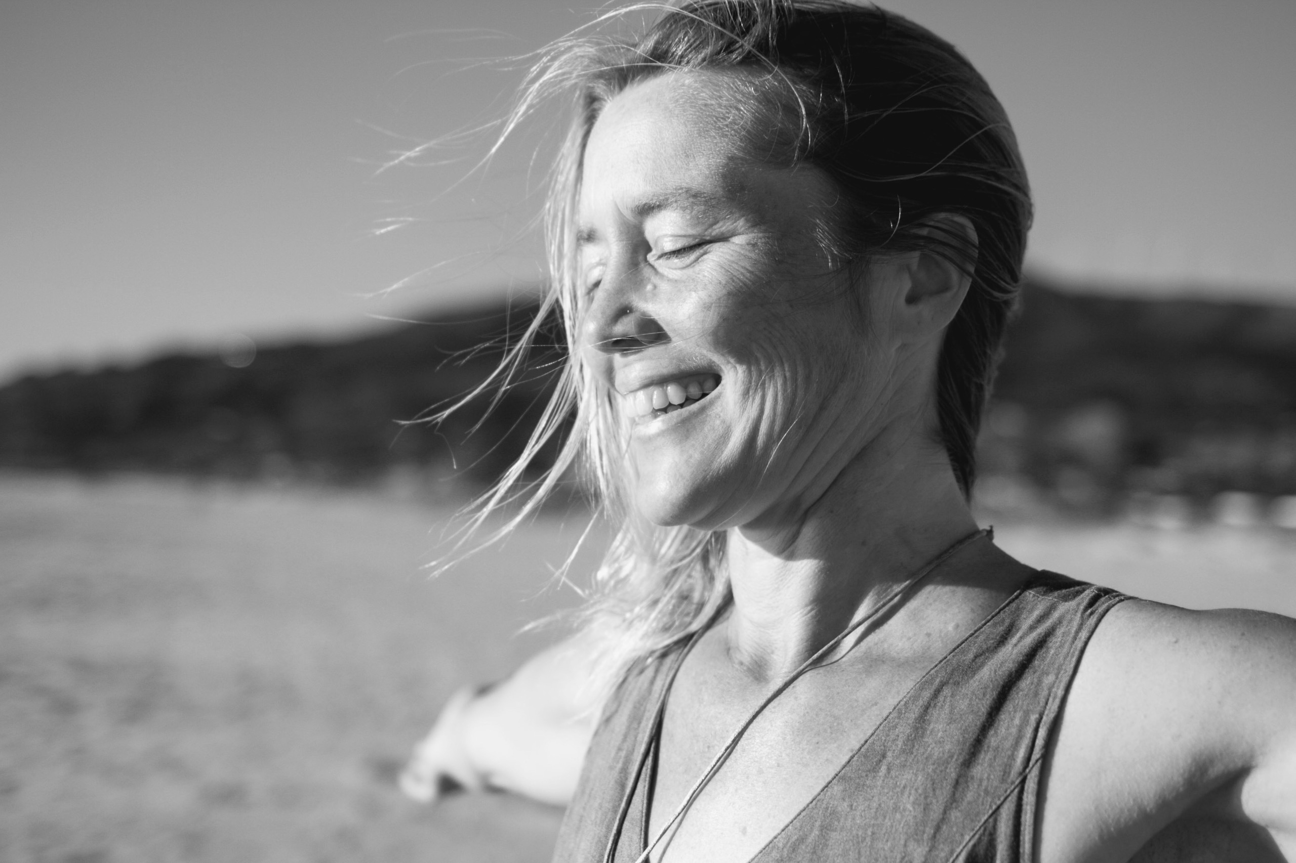 Auf dem Bild sieht man Astrid Schröder. Das Bild ist komplett in schwarz-weiß gehalten. Astrid Schröder lacht fröhlich und hat die Augen geschlossen und die Arme weit ausgebreitet. Man sieht eine wunderschöne Landschaft im Hintergrund, 